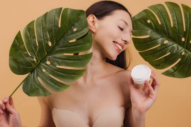 Elinde bir kavanoz kozmetik kremi ve tropikal canavar yaprakları tutan mutlu kadın, bej bir arka planda cilt için doğal güzellik ürünleri tanıtıyor..