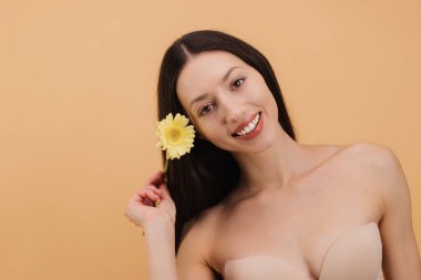 Bej arka planda sarı Gerbera çiçeği tutan gülümseyen esmer kadının stüdyo portresi.