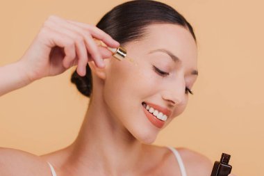 Gülümseyen genç Asyalı kadın yüz serumu kullanıyor, pipetini tutuyor, gerekli yağı sürüyor, mükemmel cildini nemlendiriyor, bej arka planda izole edilmiş.