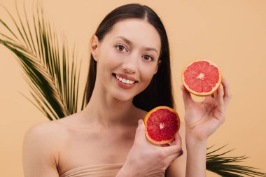 Greyfurt yarımları tutan uzun saçlı genç bir kadın yeşil yapraklı yumuşak bir arka plana gülümsüyor. Doğal güzellik ve sağlık vurgulanıyor