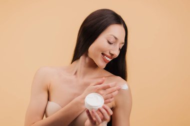 Omzuna nemlendirici krem süren mutlu genç bir kadın. Bej arka planda vücut bakımı ve cilt tedavisi kavramı..