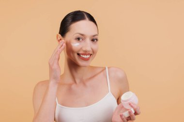Mutlu esmer kadın yüzüne nemlendirici krem sürüyor. Sağlıklı cilt tedavisi, güzellik ve bej arka plan üzerine cilt konsepti.