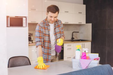Gülümseyen adam eldiven giyer mutfak masasını sprey deterjan ve mikrofiber bezle temizler sepet temizlik ürünleriyle temizler.
