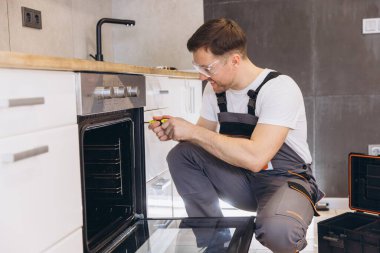 Güvenlik gözlüğü ve üniforma takan bir tesisatçı modern bir mutfak fırınını ustalıkla tamir etmek için tornavida kullanıyor.