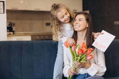 Küçük kız anneler gününü kutlayan gülümseyen annesine kırmızı laleler ve el yapımı tebrik kartı veriyor.