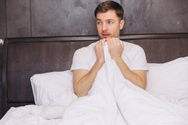 Genç adam bir battaniyenin altında saklanıyor, korkudan bunalmış ve loş bir yatak odasına endişeyle bakıyor.