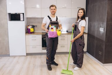 Profesyonel temizlikçiler temizlik ürünleri ve süpürge tutuyor, modern bir mutfağı temizlemeye hazır.
