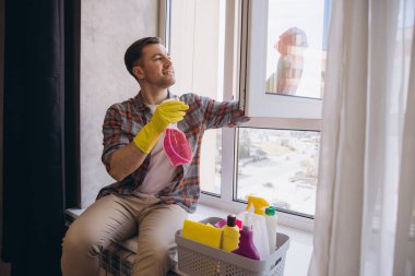 Kapıcı eldiven giyer, temizleme spreyi tutarken pencere eşiğinde oturur ve pencereleri temizler.