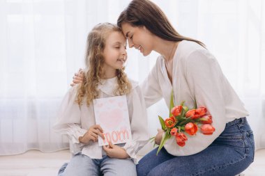 Annesine sevinçle bir tebrik kartı ve canlı kırmızı laleler sunan küçük kız, Anneler Günü 'nde sevgisini ve bağını kutluyor.