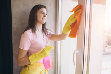 Eldiven takan, önlük giyen, pencereleri sprey ve turuncu paçavralarla temizleyen genç bir kadın.