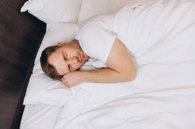 Beyaz çarşaf ve yastıklarda huzur içinde uyuyan beyaz bir adam rahat bir yatakta iyi uykuların tadını çıkarıyor.