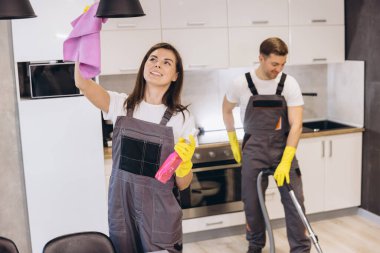 Profesyonel temizlikçiler gri üniformalar ve sarı eldivenler giyer mutfak yüzeylerini temizler ve mikrofiber bez ve sprey şişeyle lamba taşırlar.