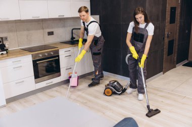 Temizlik görevlileri mutfak zeminini paspaslıyor ve süpürüyor, temiz ve hijyenik bir ortam sağlarken ev işleriyle uğraşıyor.