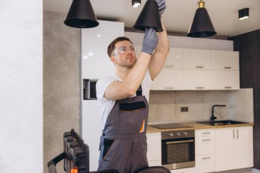 Güvenlik gözlüğü ve eldiven takan elektrikçi çağdaş bir mutfağa modern bir kolye lambası yerleştiriyor.