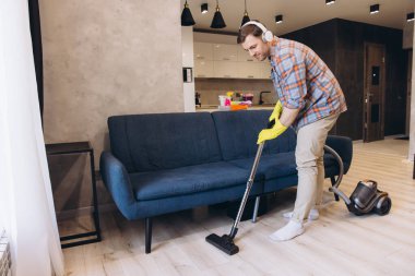 Kulaklık takan ve sarı eldiven takan genç bir adam modern bir dairede mavi bir kanepenin yanında yerleri süpürüyor.