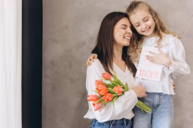 Kızı annesine sarılıyor ve Anneler Günü için ona bir tebrik kartı ve bir buket lale veriyor.