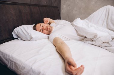 Genç adam rahat bir yatakta beyaz çarşaflarla uyur, iyi bir uyku ve rahatlığın tadını çıkarır.