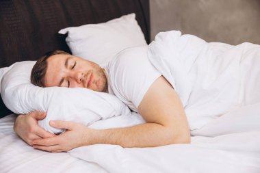Kafkasyalı bir adam, yumuşak beyaz çarşaflarda huzur içinde uyuyor, pelüş bir yastığı kucaklıyor, sakin ve rahatlatıcı bir anın tadını çıkarıyor.