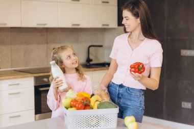 Anne ve kızı gülümserken, modern mutfakta taze yiyecek paketleri açıyorlar, sağlıklı beslenme alışkanlıklarını ve aile zamanlarını destekliyorlar.