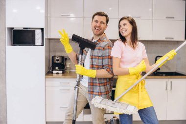 Gülümseyen çift lastik eldiven takıyor süpürge ve süpürge kullanıyor, birlikte mutfağı temizliyor ve eğleniyorlar.
