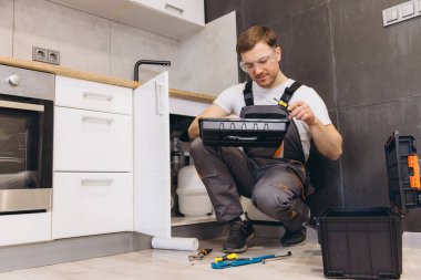 Profesyonel tesisatçı, mutfak lavabosunun altında özenle çalışıyor, çeşitli aletler kullanıyor ve güvenlik gözlüğü takıyor.