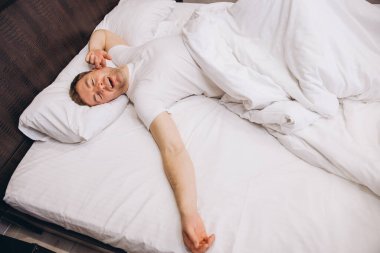 Yatakta huzur içinde uyuyan bir adam, uyandıktan sonra esniyor ve esniyor, yumuşak beyaz çarşaflara sarılıyor, rahat bir sabah anının tadını çıkarıyor.