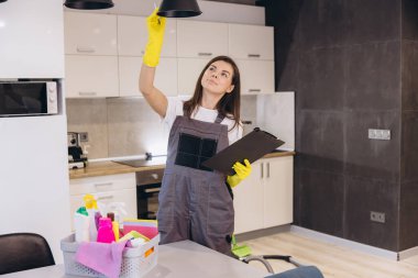 Üniformalı ve sarı eldivenli profesyonel temizlikçi kadın, modern bir mutfakta lambayı kontrol ediyor, elinde bir pano tutuyor.