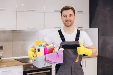 Profesyonel temizlikçi elinde temizlik ürünleri olan bir sepet tutuyor. Başparmaklarını gösteriyor, modern bir mutfağı temizlemeye hazır.