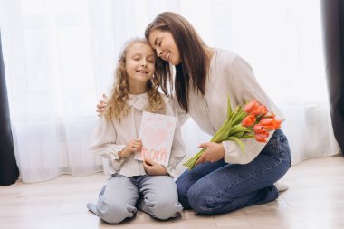 Kızı annesine tebrik kartı ve lale veriyor, sarılıyor ve gülümsüyor, Anneler Günü 'nü kutluyor.