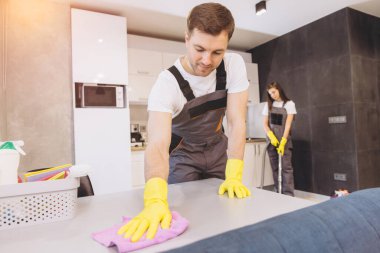 Gri üniformalar ve sarı eldivenler giyen temizlik şirketi çalışanları mutfak masasını ve zemin temizliyor.