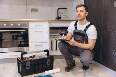 Tesisatçı çömeliyor, mutfak lavabosunun altındaki boruları inceliyor ve yanında alet çantasıyla panoya not alıyor.