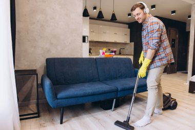 Genç bir adam modern bir oturma odasının ahşap zeminini süpürürken kulaklıkla müzik dinliyor, günlük temizlik rutininin keyfini çıkarıyor.