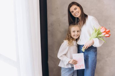 Gülümseyen annesine neşeli bir tebrik kartı ve canlı laleler sunan kız birlikte Anneler Günü 'nün neşesini kutluyorlar.