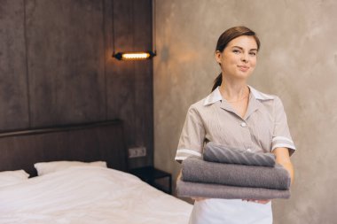 Otel odasında temiz havlu tutan genç oda hizmetçisi misafirler için onları yatağa yatırmaya hazır.