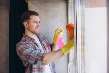 Genç gülümseyen erkek hademe eldiven giyip cam camını deterjan spreyi ve turuncu bezle temizliyor.