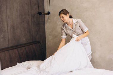 Genç oda hizmetçisi otel odasında çarşaf değiştiriyor. Misafirler için temiz ve rahat bir ortam sağlıyor.