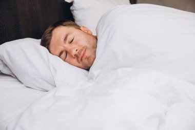 Genç adam yatakta dinleniyor, beyaz çarşafların altında huzurlu ve rahat bir uyku çekiyor.