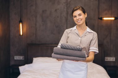Genç oda hizmetçisi gülümserken elinde yeni katlanmış havlularla modern bir otel odasında üniformasında sıcaklığı ve konukseverliği somutlaştırıyor.