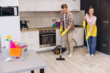 Genç, gülümseyen çift elektrikli süpürge ve paspas kullanarak lastik eldiven takıyor, modern apartmanda mutfak zeminini birlikte temizliyor.