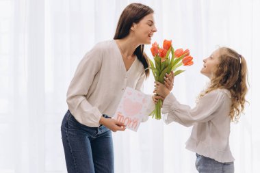 Küçük kız, Anneler Günü 'nde gülümseyen annesine kırmızı lale ve tebrik kartı veriyor.