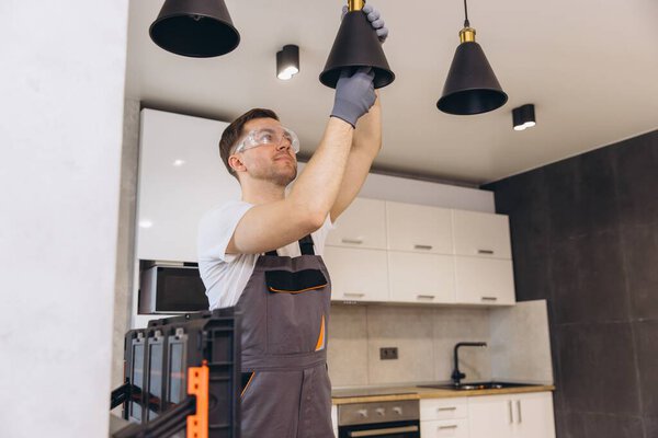Профессиональный электрик, работающий с защитными очками и перчатками, устанавливающий подвесную лампу на современной кухне