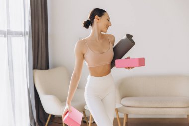 Yoga hocası yoga minderi ve bloklar taşırken gülümsüyor. Parlak oturma odasında online dersi için hazırlanıyor.