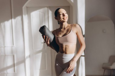 Elinde yoga minderi tutan sağlıklı bir kadın, egzersiz rutinine hazır, içeride sağlık ve sağlığı destekliyor.