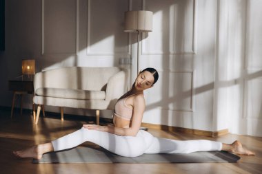 Genç bir kadın evdeki yoga minderinde bacaklarını ayırarak günlük esneme ve farkındalık rutininin tadını çıkarıyor.