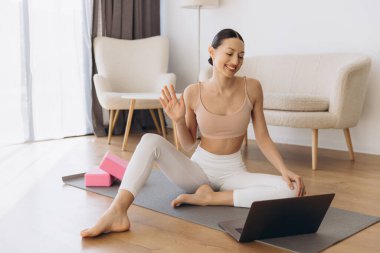 Yoga hocası öğrencilerine neşeyle el sallarken aynı zamanda şık oturma odasından online bir ders veriyor.