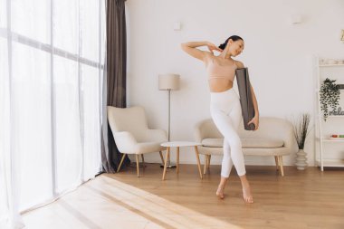 Spor kıyafetleri giyen, yoga minderi tutan, modern, parlak oturma odasında çıplak ayakla yürüyen, ahşap zemini ve mobilyaları olan sağlıklı bir kadın.