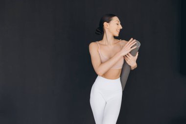 Yoga hocası elinde yoga minderi tutarken gülümsüyor, evde yoga kursuna hazırlanıyor.