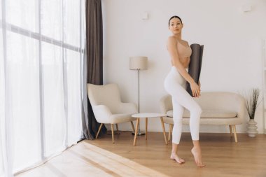 Elinde yoga minderiyle dingin bir oturma odasında egzersiz yapmaya hazırlanan sağlıklı bir yaşam tarzını ve sağlığını kucaklayan sağlıklı bir kadın.