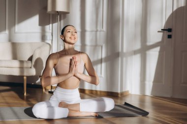 Lotus pozunda meditasyon yapan bir kadın, evde minder üzerinde yoga yapan, pencereden gün ışığının tadını çıkaran bir kadın.