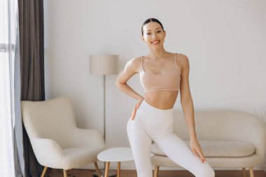 Spor kadın evde yoga yapıyor, spor giysiler giyiyor, oturma odasında minimalist mobilyalarla duruyor.
